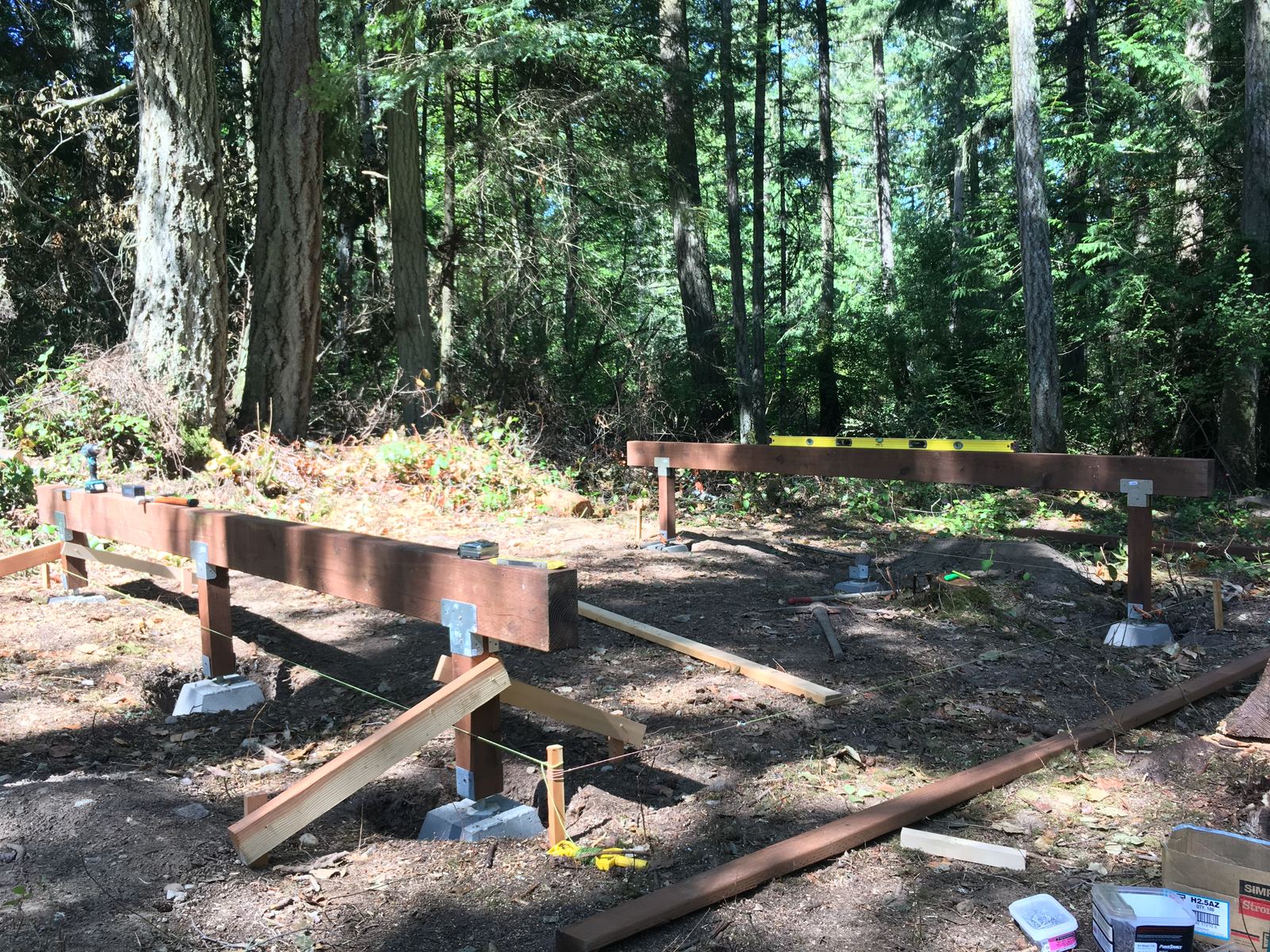 Two long stained beams set on precast concrete piers with steel post bases, in a patch of second-growth forest cleared down to bare dirt. A yellow spirit level lies along the far beam, mason's string runs between stakes, and a box of framing ties sits in the corner of the frame.