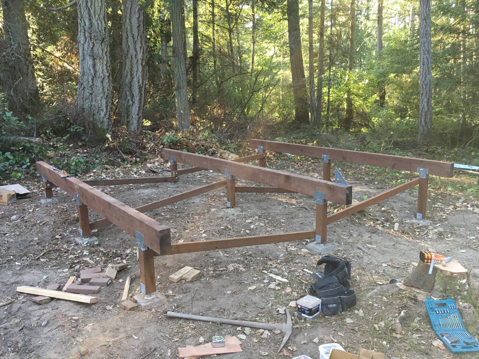 The pier-and-beam foundation further along: a rectangle of stained beams standing on concrete piers, with diagonal braces run down to the posts. A tool belt, a mattock, a drill bit case and a speed square lie about on the forest floor.