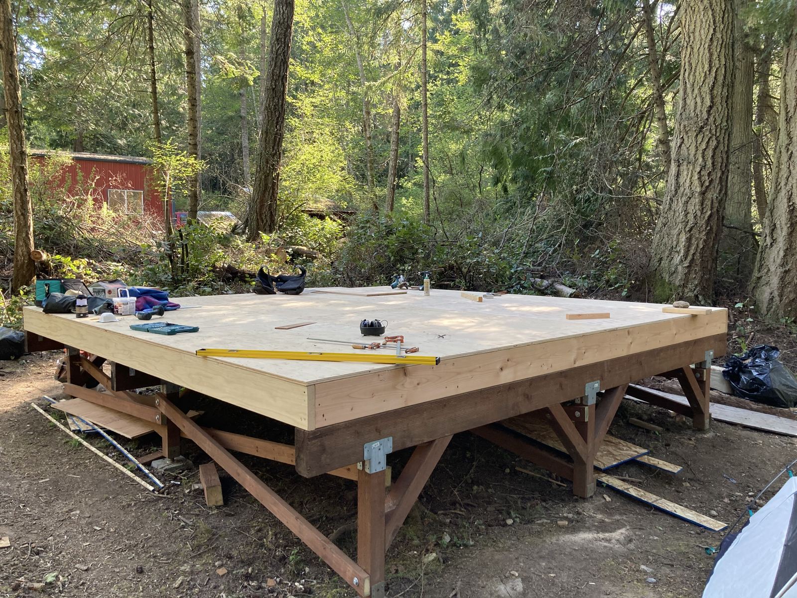 The finished floor platform, decked in plywood and standing waist-high on stained posts with knee braces at every corner. A yellow level, a hard hat, a chalk line and a drill lie on the deck; a red shed and a parked car sit back in the trees.