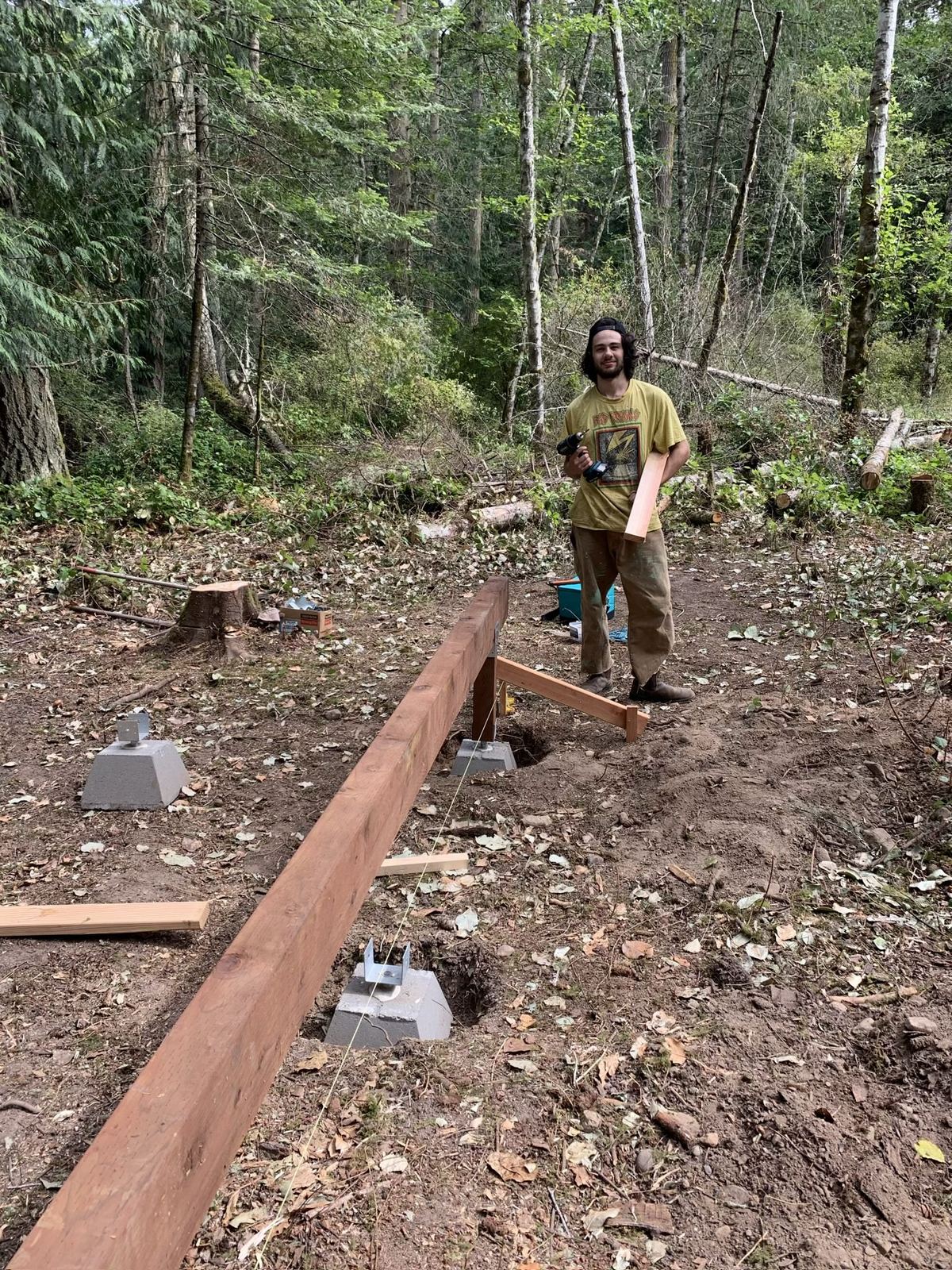 A man in a yellow t-shirt and work trousers standing in the cleared site with a drill in one hand and a length of framing lumber under his arm, grinning at the camera. A stained beam runs across the foreground on its pier, string lines pulled taut beside it.
