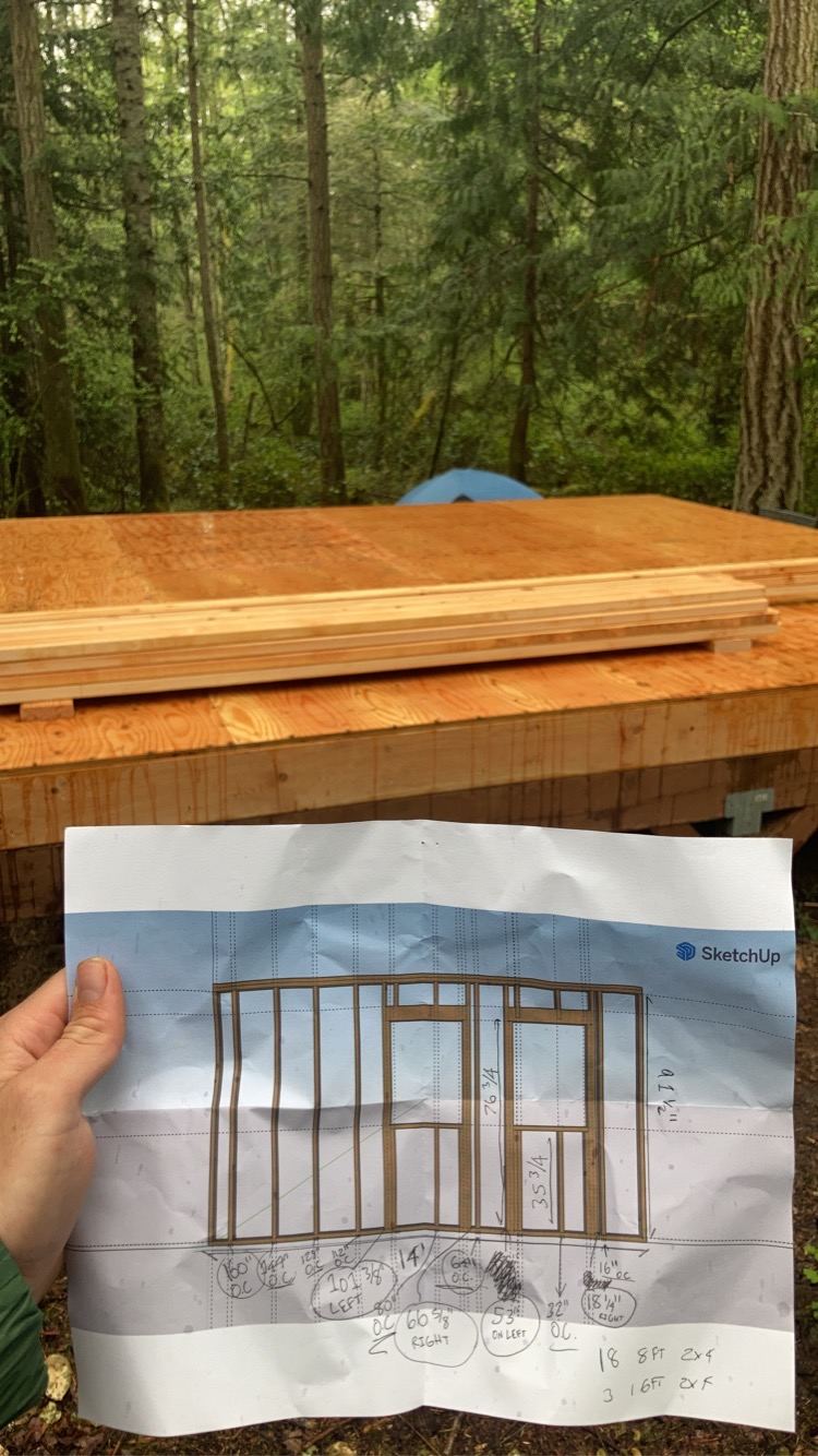 A hand holding up a printed SketchUp elevation of the wall framing, marked over in pencil with stud spacings, rough opening sizes and a cut list at the bottom reading 18 8ft 2x4 and 3 16ft 2x4. Behind it the finished floor platform stacked with framing lumber, and a blue tent in the trees.