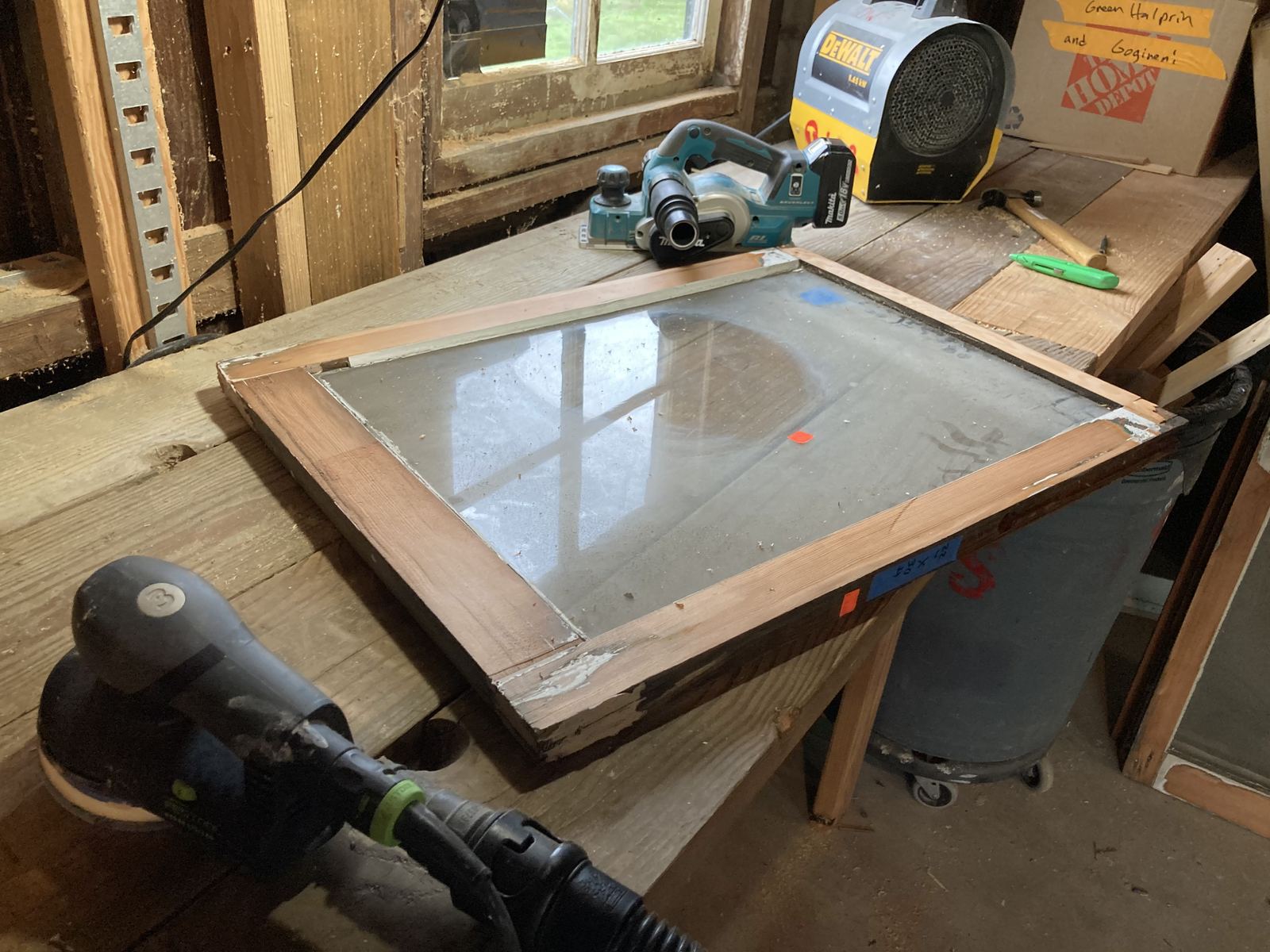 A salvaged window sash on a workbench with the glass masked in blue tape, alongside a cordless planer, a random orbit sander on dust extraction, a hammer and a marking pencil. A cardboard box behind is labelled in marker.