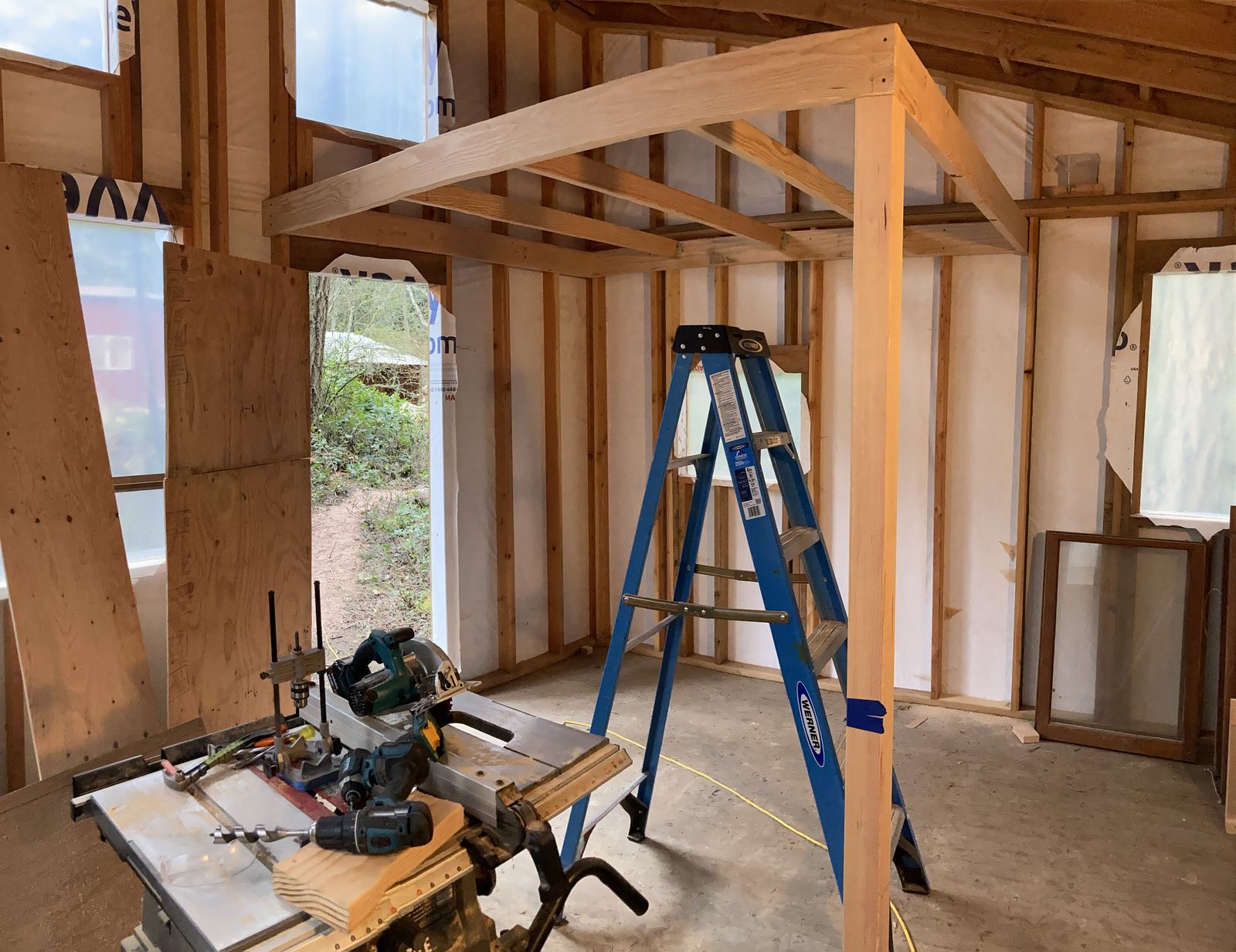 Inside the shell: the loft header framed in and carried on a post, studs and housewrap all round, a blue step ladder underneath. A mitre saw and a drill press stand on a bench in the foreground and salvaged window sashes lean against the wall.