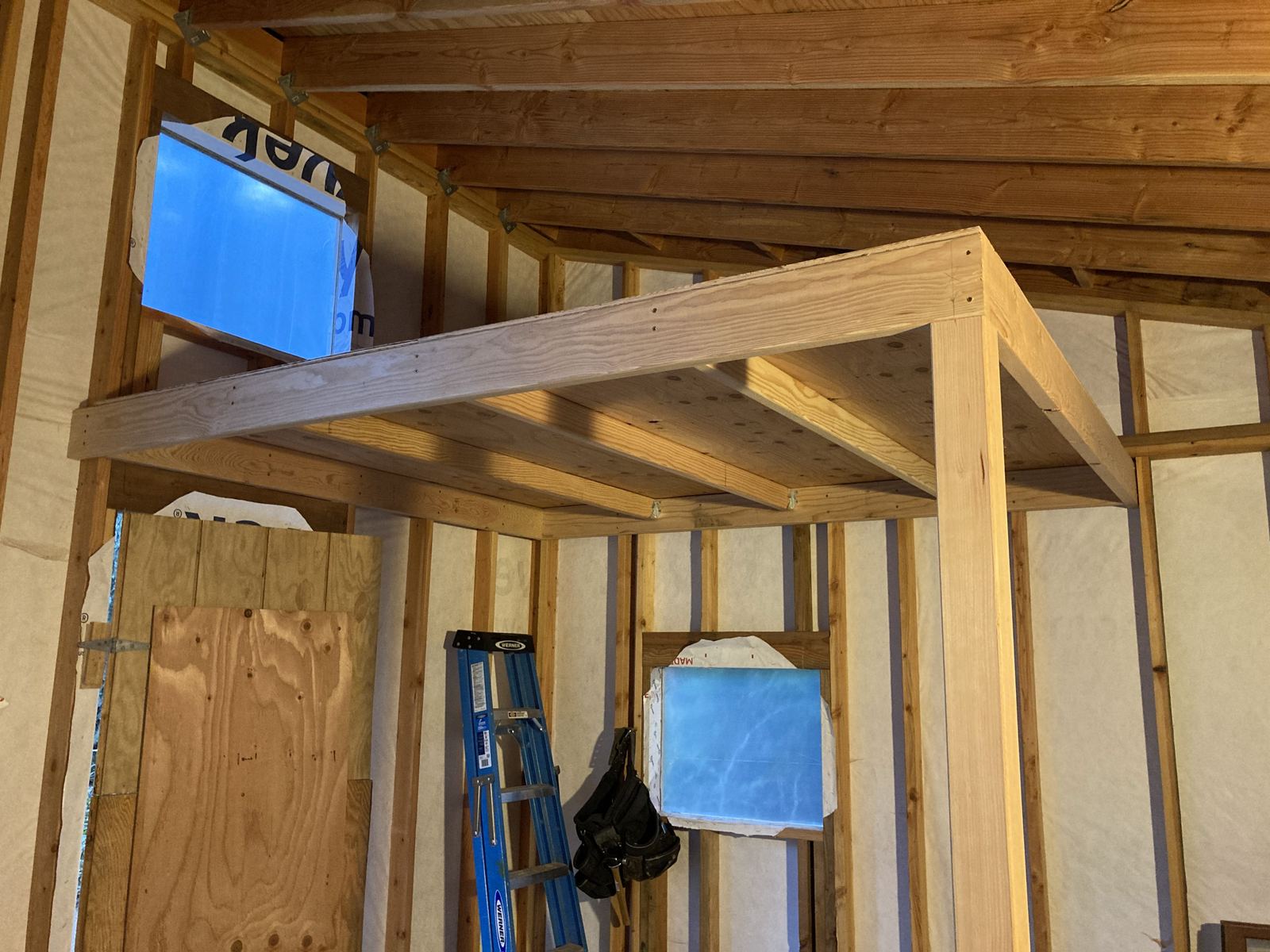 The loft platform framed overhead, joists let into a header on a post with plywood laid on top. A small square window at the far end and a tool belt hanging from a stud.