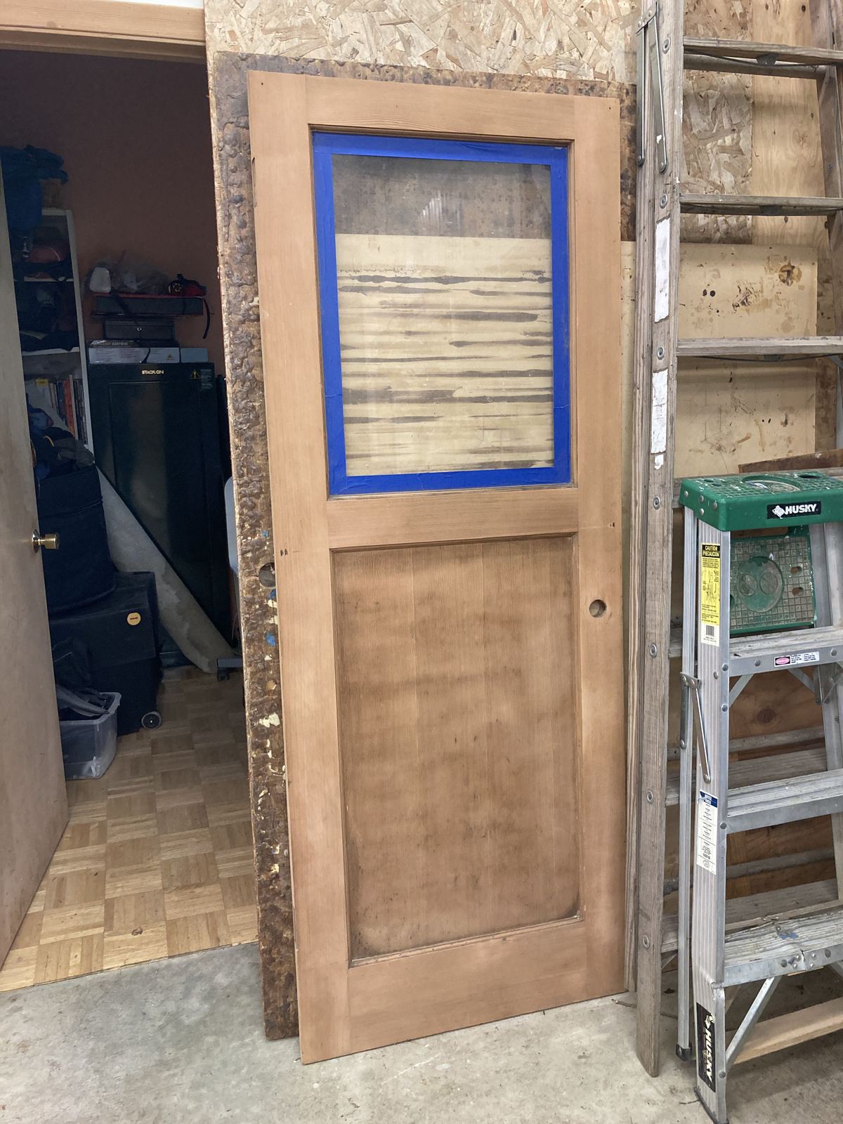 A salvaged fir door standing against a garage wall, stripped back to bare wood with the old finish still showing in streaks, its upper glass panel masked in blue tape. A ladder and a tool tray stand beside it.