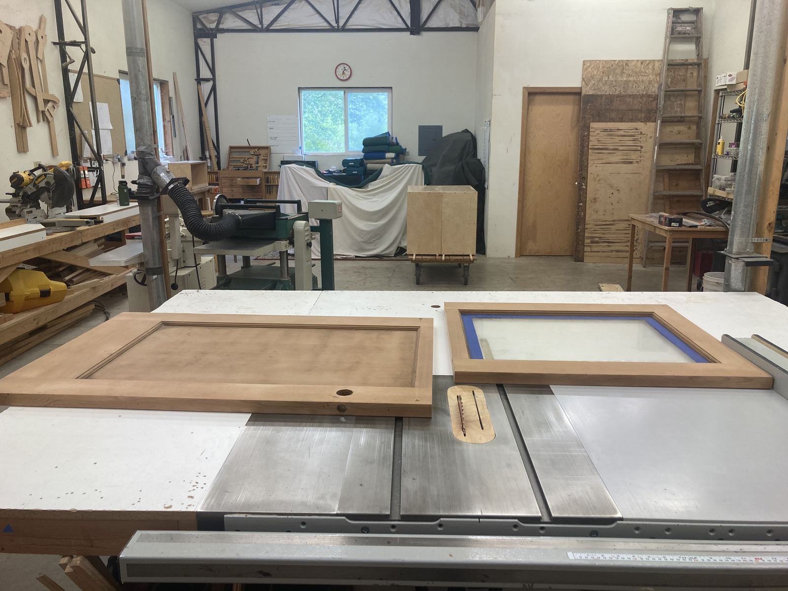 The same door and a window sash laid out on a table saw in a woodshop, both stripped and sanded, the glass taped. Behind them a mitre saw, dust extraction, a bandsaw, plywood stacked against the wall and a clock above a window.