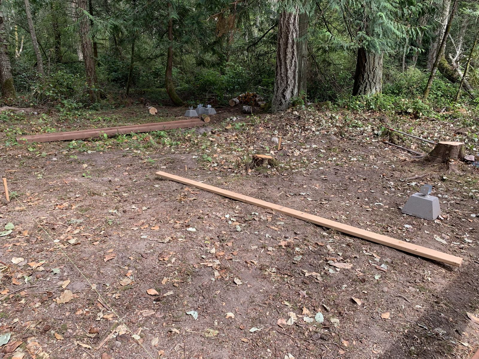 The site nearly bare: string lines pegged out across the dirt, a stained beam laid along one edge, a fresh length of framing lumber across the middle, and concrete piers with steel bases set and waiting.