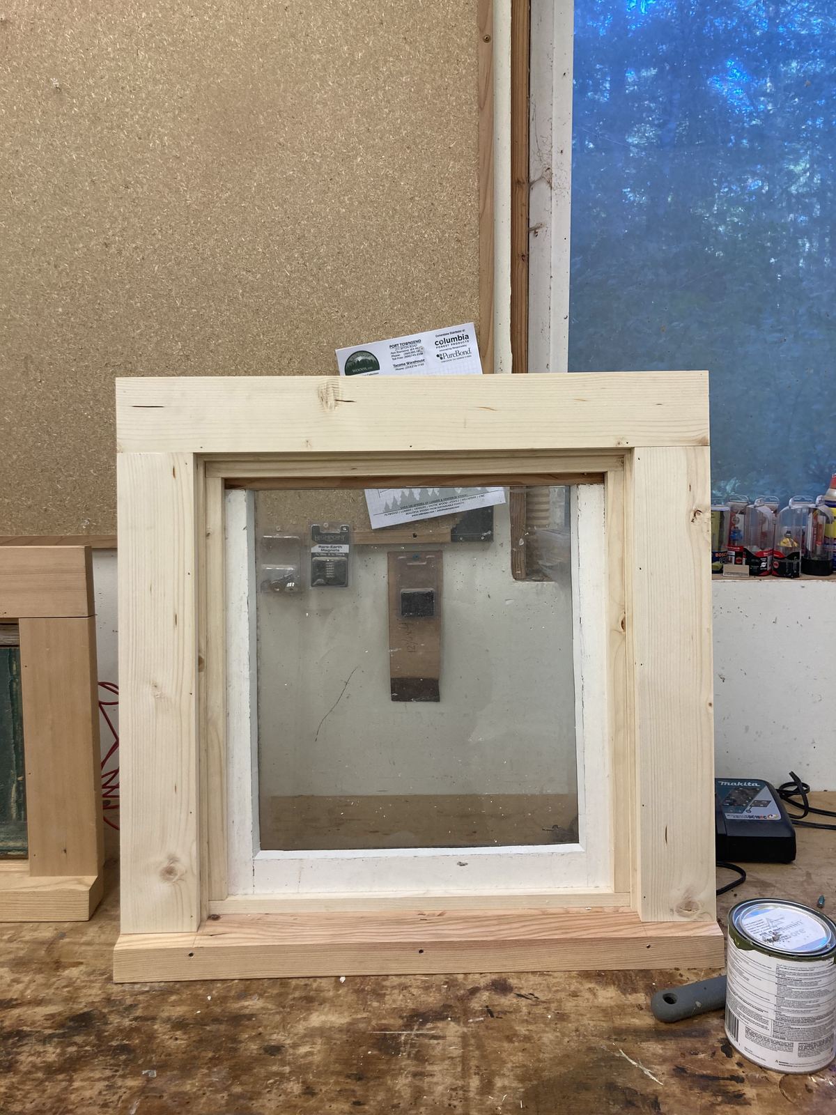 A window fitted into a new frame with casing and a sill, standing on a workbench against an OSB wall, the glass still dusty from the shop. A battery charger and a tin of paint sit to the right.