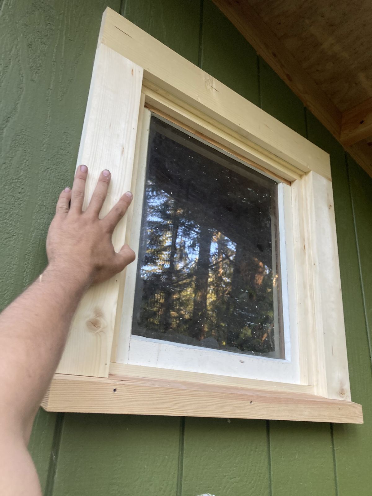 A hand holding the finished window frame up against the green siding under the eave, checking how it sits. The trees behind the camera show in the glass.