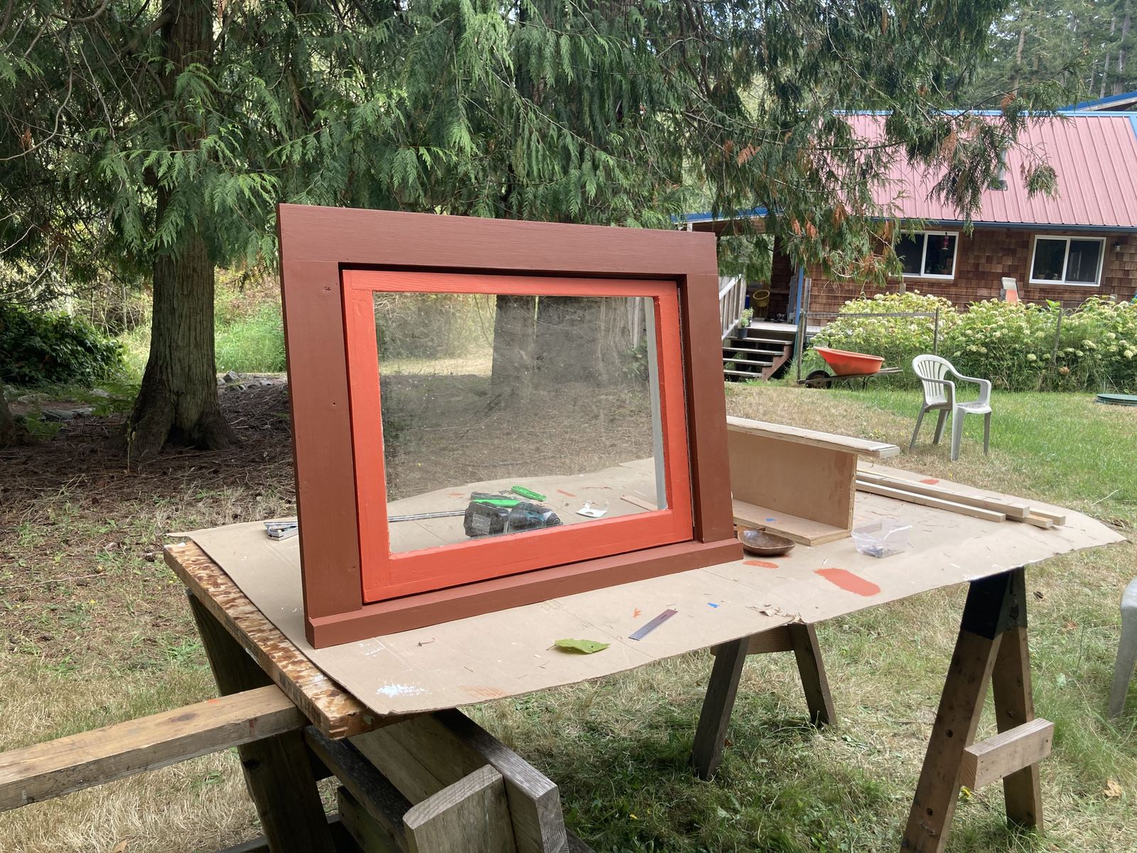 The window casing painted dark red with a bright orange inner bead, standing on cardboard over sawhorses in a mown yard. A cedar-shingled house with a red metal roof sits in the background.