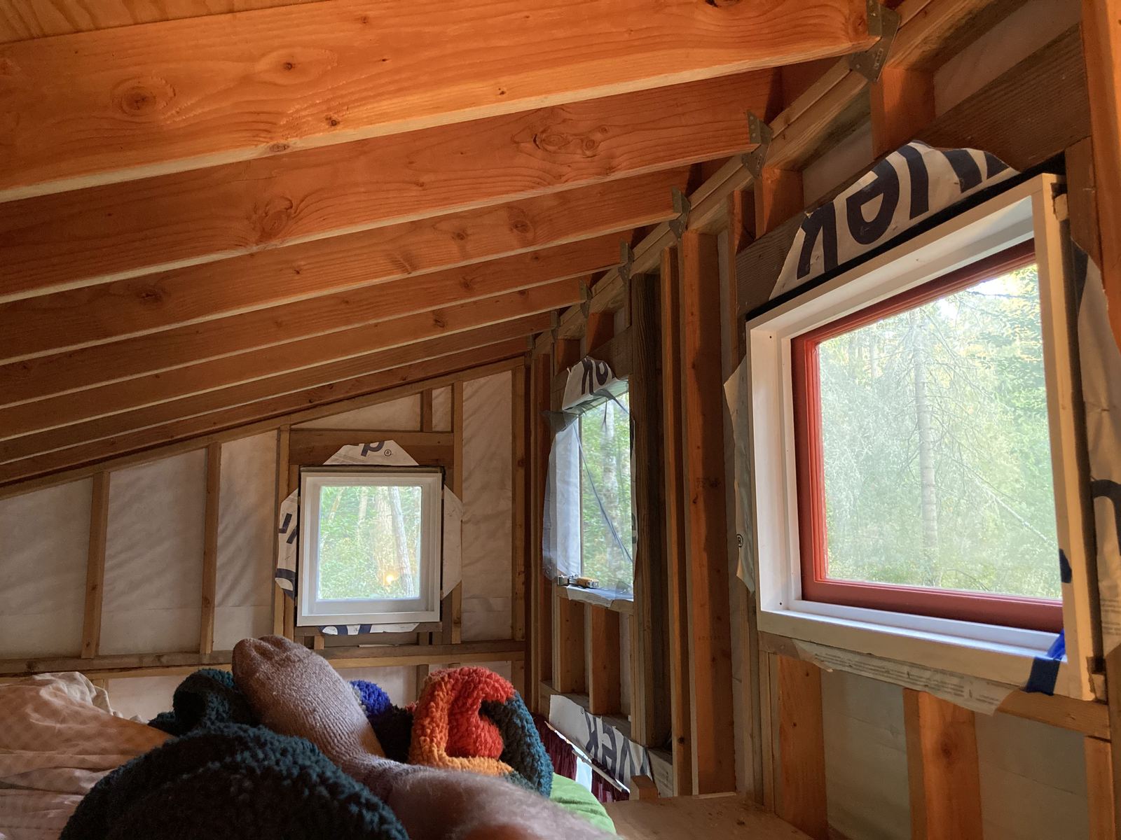 Inside the loft, looking along the sloping ceiling of exposed rafters and plywood. Three windows in the wall, one small and square, one open, one cased in orange, with housewrap between the studs and bedding in the foreground.