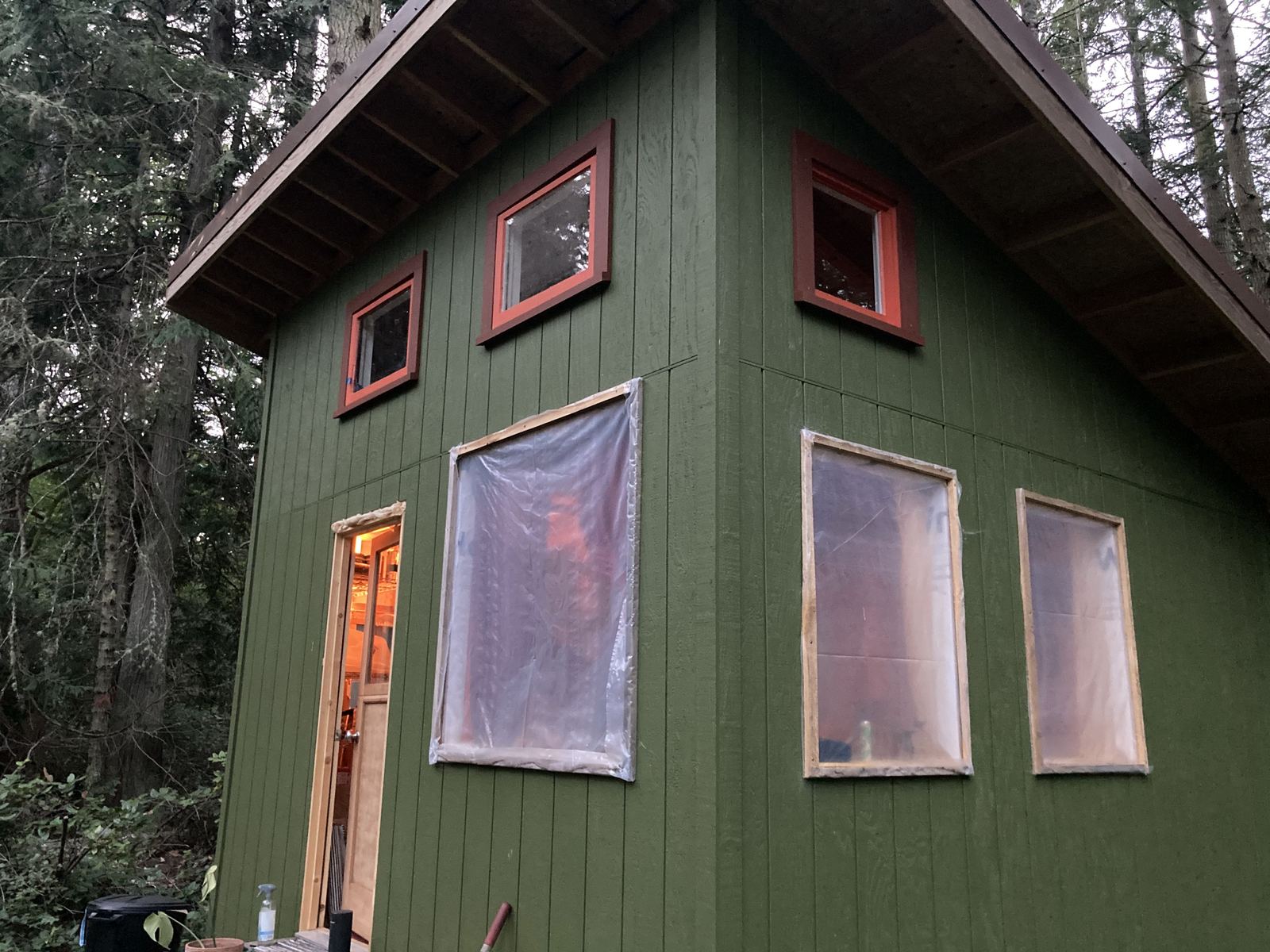 The cabin at dusk from the corner: green siding, four small windows cased in red, the larger openings still covered with taped plastic sheet, and the door standing open with warm light inside.