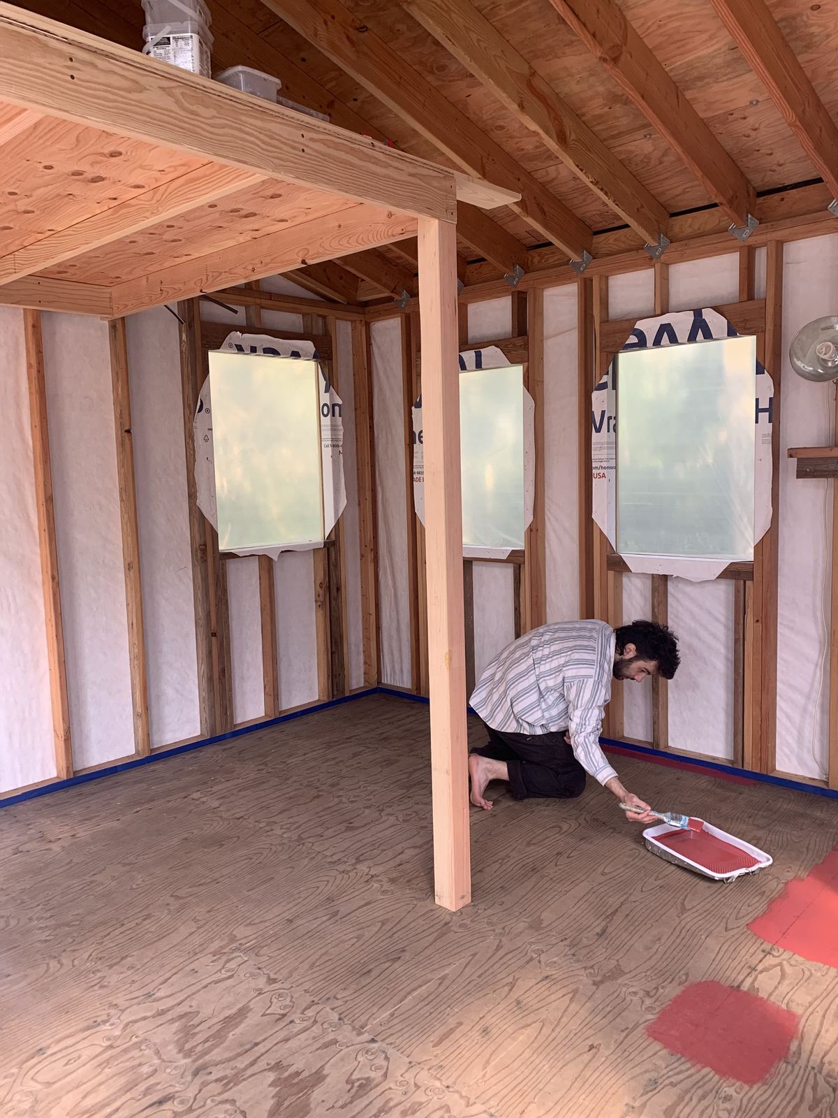 Inside, a man kneeling barefoot to paint the plywood floor red with a brush and a roller tray. The walls are framed and wrapped, three windows are in, and the loft header runs overhead on its post.