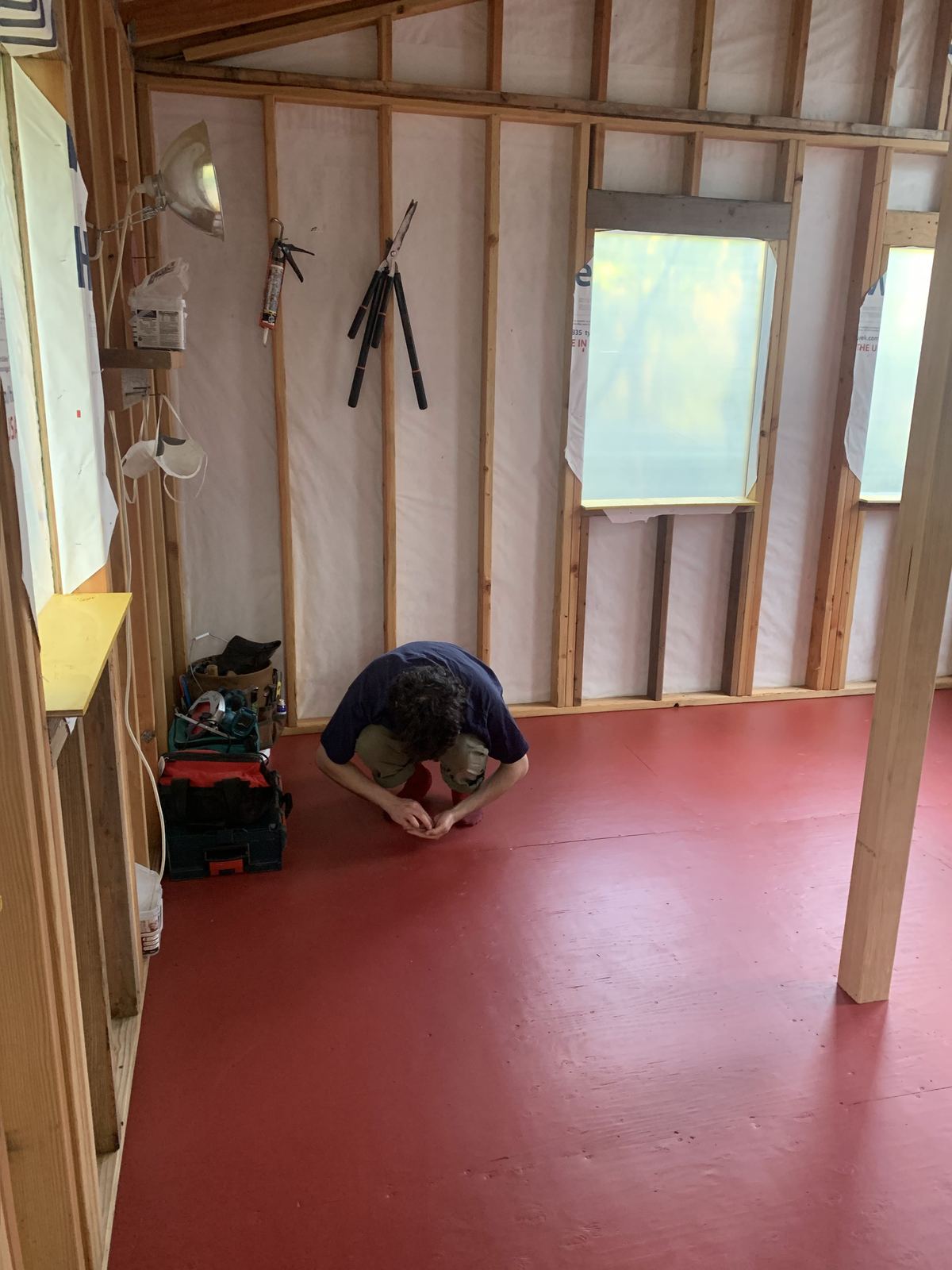 Inside again with the floor finished in bright red, the man crouched over something on it. A work light, a caulk gun, loppers and a dust mask hang from the studs, and tool bags are stacked in the corner.