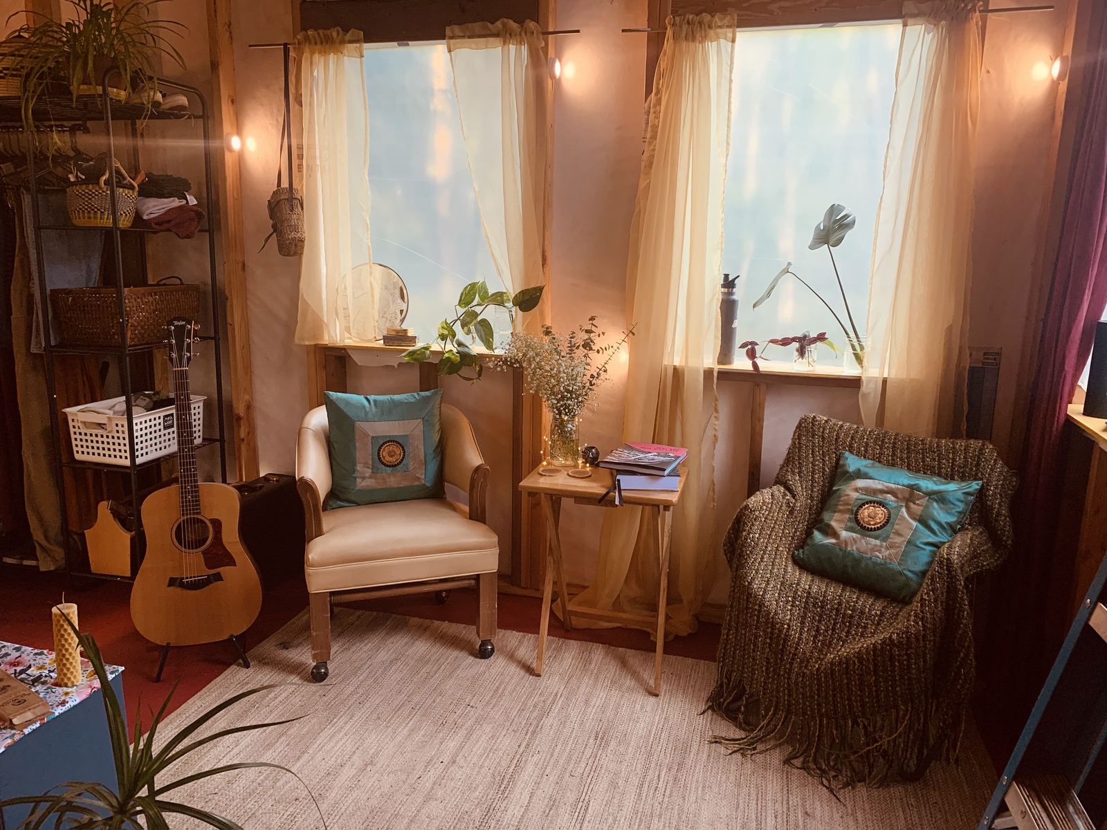 The finished interior: two curtained windows, string lights along the wall, an acoustic guitar leaning against a shelving unit hung with clothes and baskets, a leather armchair, a wicker chair under a throw, and a folding table with books and a jar of dried flowers.