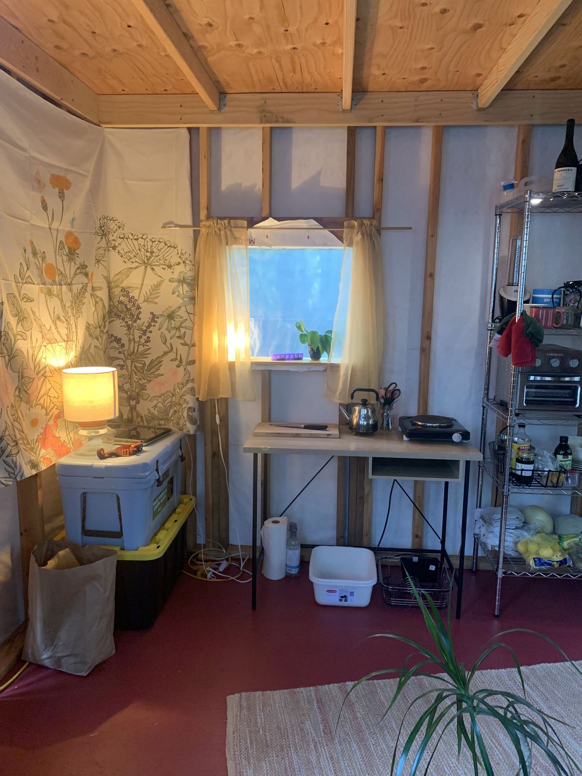 A corner set up as a kitchen: a work table with a kettle, a hot plate, a knife and board, a curtained window above it, a cooler and bins along the wall, and a botanical print cloth hung as a wall covering.