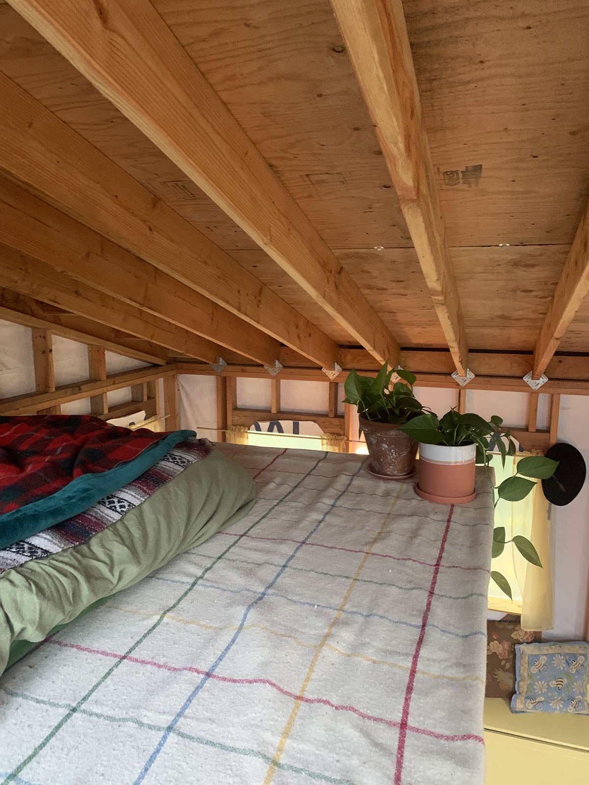 The loft made up as a bed under the sloping plywood ceiling, a woven blanket in coloured checks with quilts folded at the head, two pothos in pots on the ledge and a hat hung on a nail.