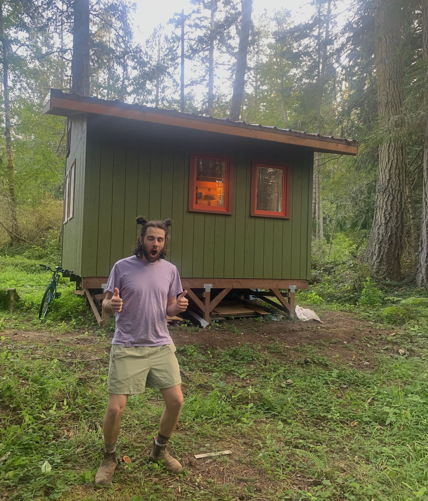 The finished cabin from the front, olive green with red window casings and a metal shed roof, standing on its post foundation in the woods. A man in a purple t-shirt and shorts stands in the grass giving two thumbs up, mouth open mid-shout. A bicycle leans against the far corner.