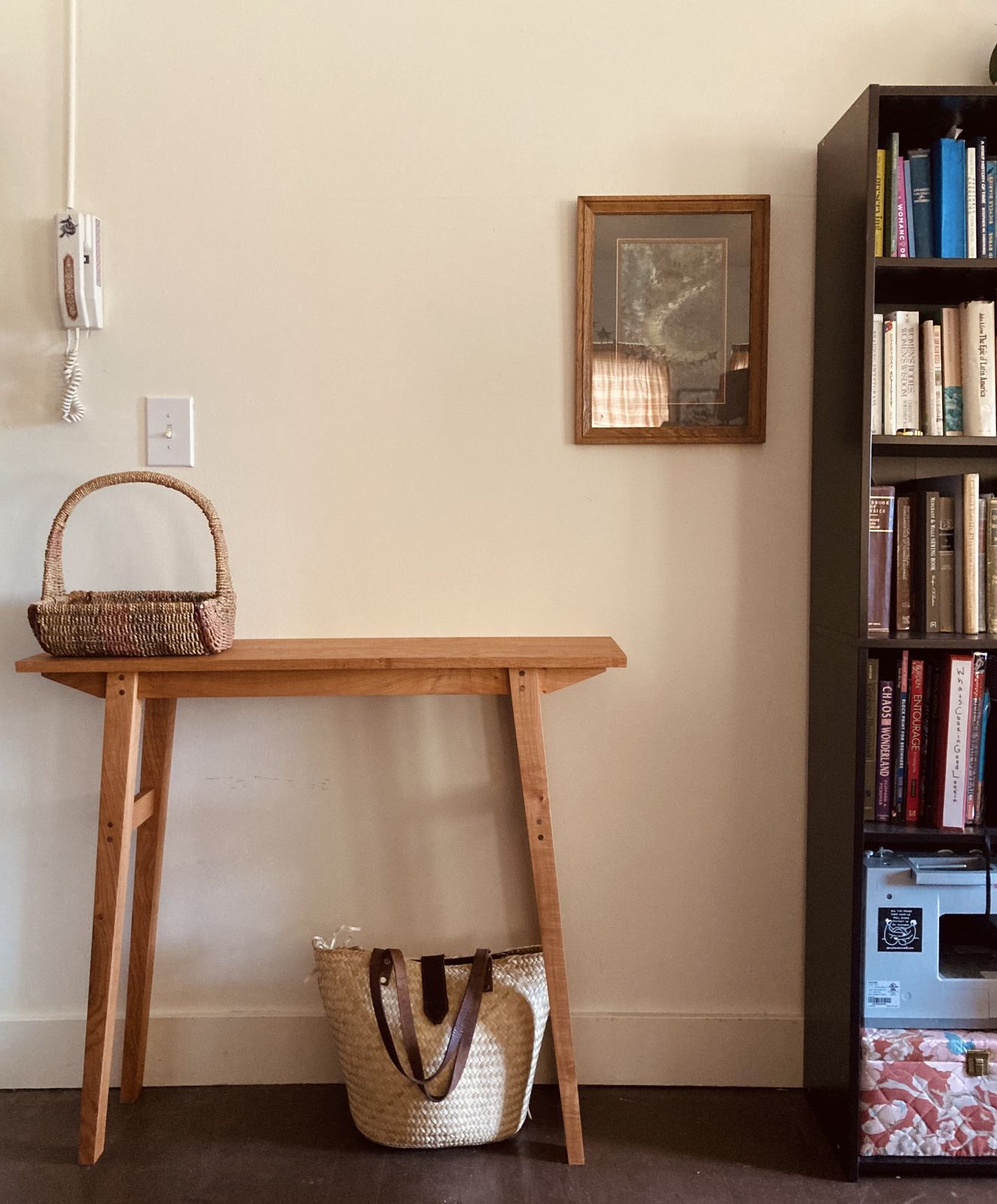 The finished table against a cream wall: a narrow cherry console with a thin top and splayed, tapered legs, pinned at the joints with visible pegs. A woven basket sits on top, a straw tote underneath, a framed picture and a bookshelf to the right.