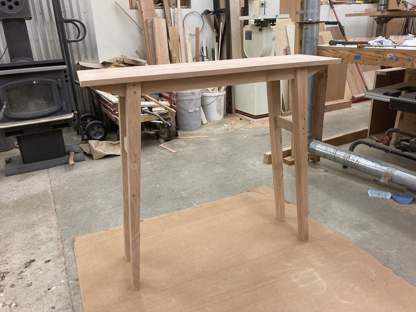 The same table unfinished, standing on cardboard in the shop. Bare cherry, pegs showing at every joint, the underside stretcher visible. Behind it a bandsaw, a woodstove, buckets, offcuts and a bench.