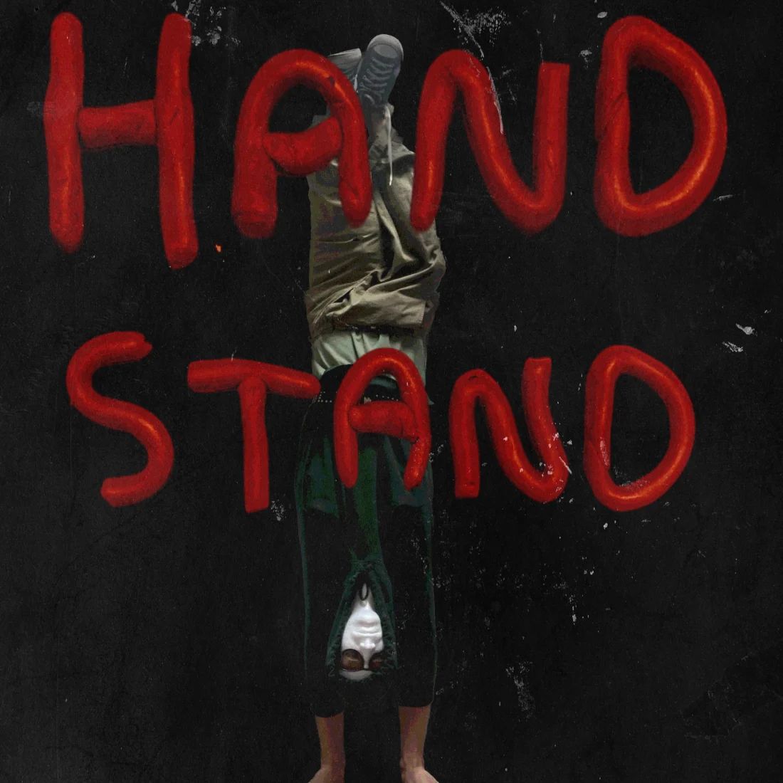 A figure upside down in a handstand against a scuffed black wall, bare feet at the bottom of the frame and sneakers at the top, shirt hanging down over an inverted face. HAND STAND is daubed across the wall in thick red paint, the word split over two lines.
