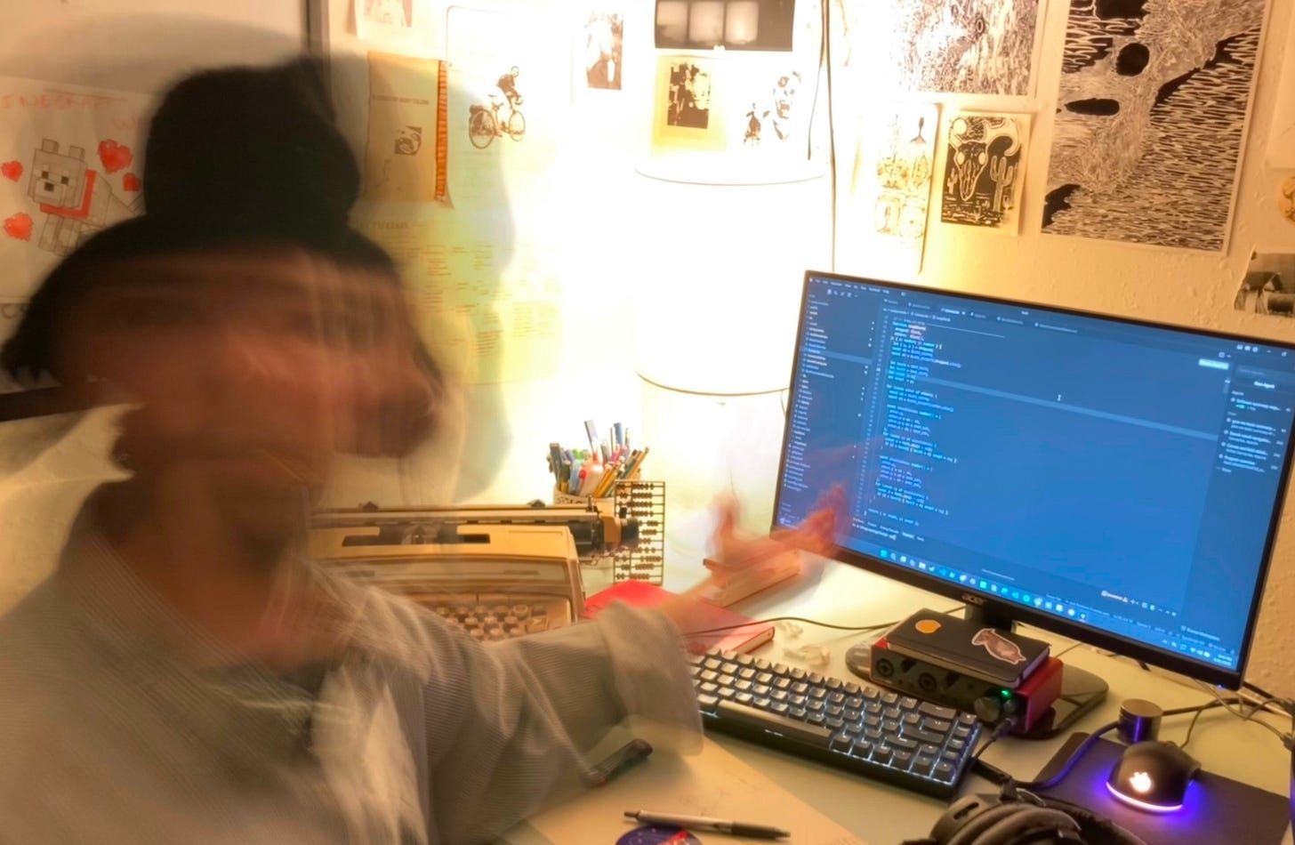 A blurred figure at a desk at night, moving fast enough that the head and one arm smear across the frame. On the desk a mechanical keyboard, a wide monitor filled with code, a typewriter pushed to the back, a jar of pens, a small red box of electronics, headphones and a mouse. The wall behind is papered edge to edge with drawings, prints and photographs.
