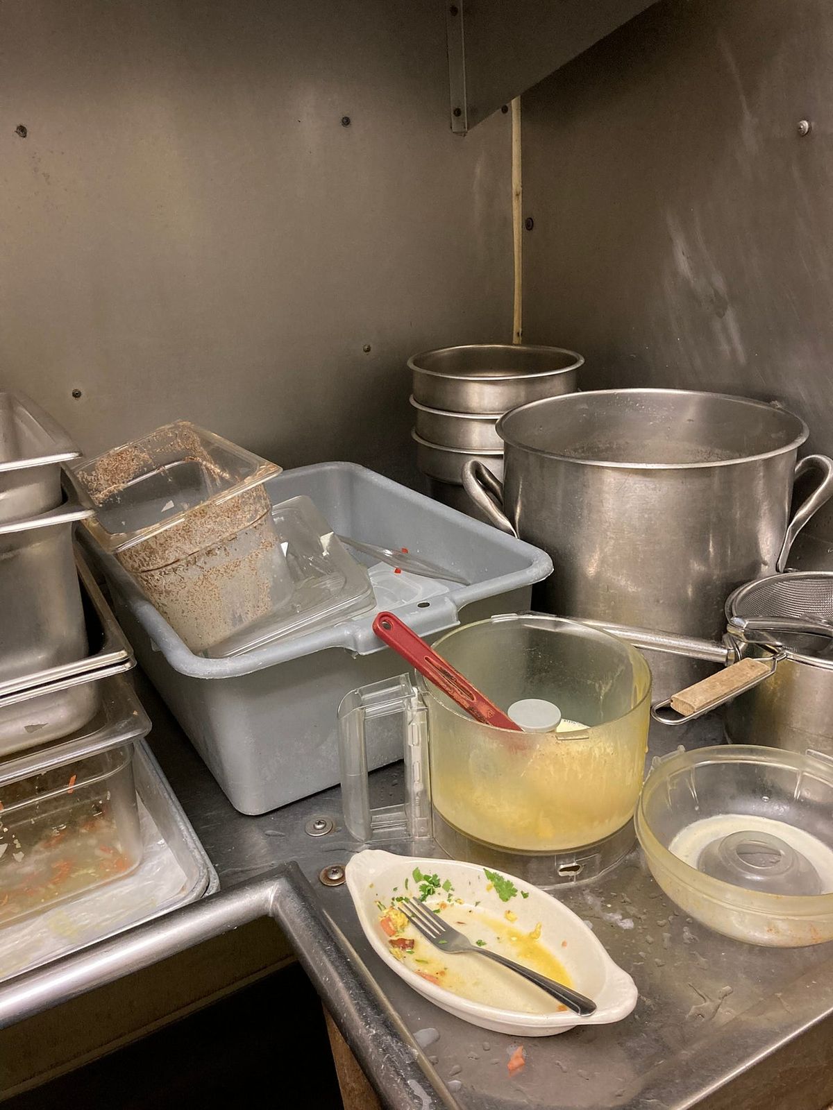 A commercial kitchen dish pit partway through a shift. On a stainless counter, a grey bus tub holds cloudy plastic containers crusted brown; behind it a tall stockpot, a stack of round pans, a conical strainer with a wooden handle. In front, a food processor bowl smeared yellow with a red spatula standing in it, and a white oval plate with a fork laid across it, orange sauce and bits of parsley left behind.