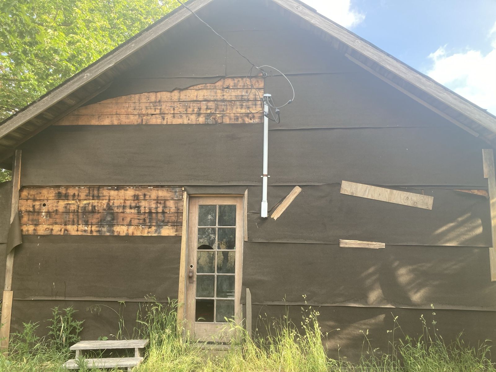 The gable end of an old house with its shingles stripped off and black tar paper over most of the wall, bare weathered boards showing through in two long patches. A single glazed door with nine panes stands at the centre with a wooden step in the grass in front of it. A white electrical mast is bolted beside the door with wire looping off it. Tall grass across the foreground, a maple at the left, blue sky at the right.