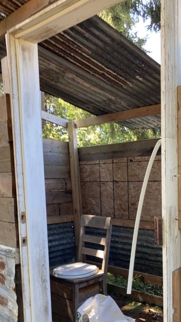 Inside a small outbuilding, a wooden ladder-back chair with a white toilet seat screwed to it, standing on a plank floor over a boxed-in base. Corrugated metal roofing overhead, corrugated metal and bare OSB for walls, the far side open to the trees. A white pipe curves down through the frame.
