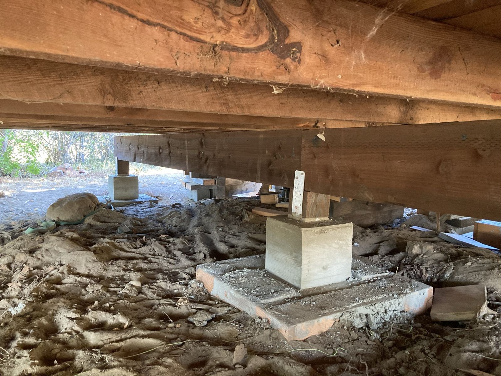 Underneath the raised house, looking along the new beams. A fresh concrete pier with a steel bracket sits on a poured pad in the foreground and another stands further off, daylight showing under the far edge. Bare dug earth below, cobwebs on the timber above.