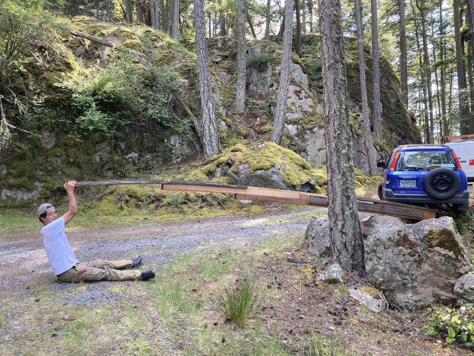 A man in a white t-shirt and cap sits on a gravel drive with his legs straight out, both hands overhead gripping the end of a long lever built from lumber lagged together. The far end of the lever runs up under a blue Honda CR-V that is hung up at the edge of the drive with a wheel off the ground. Mossy rock and fir trees behind.