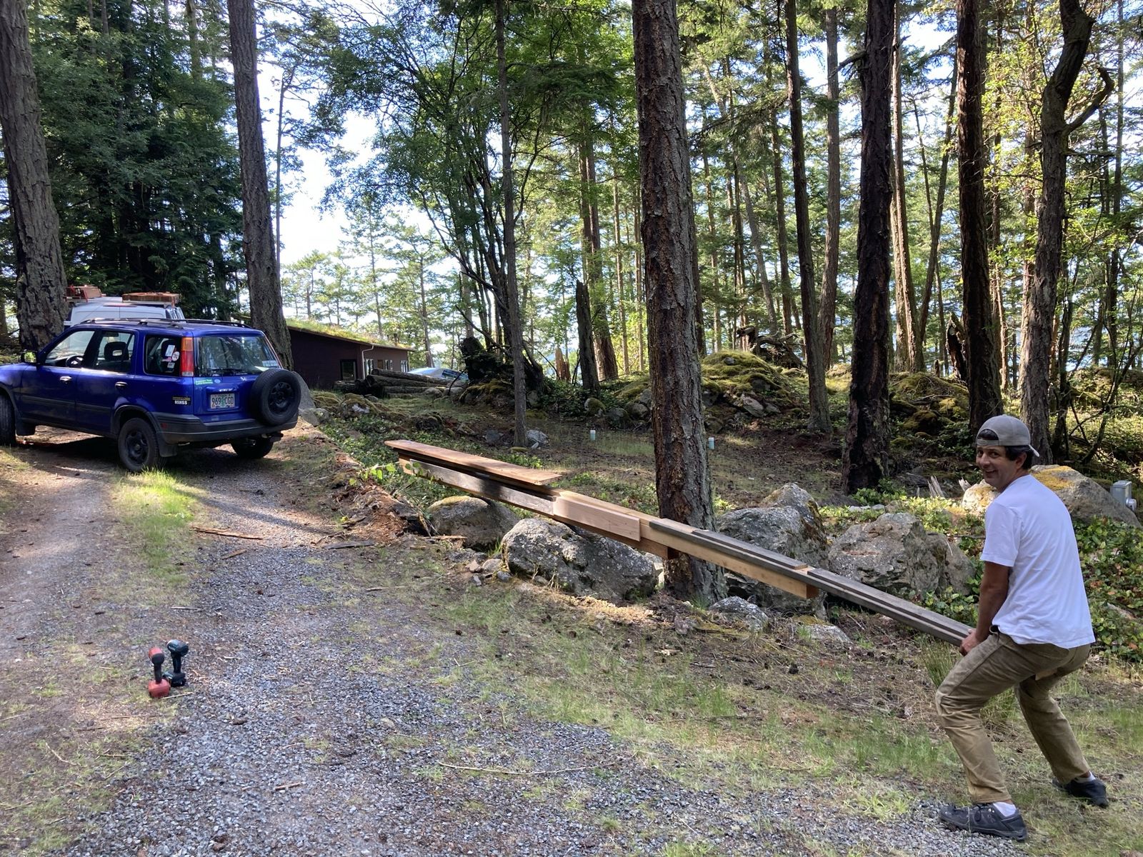The same lever from the other side, running the length of the frame. A man in a white t-shirt and cap stands at the near end, bent over it, looking back at the camera and grinning. The blue CR-V sits at the top of the gravel drive at the left with a spare wheel on the back. Two cordless drills lie on the gravel, fir forest and a glimpse of water beyond.
