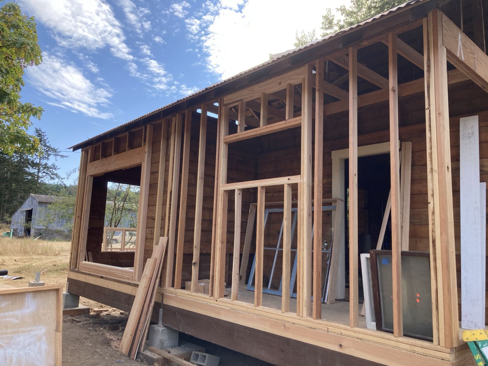 A building on new foundations with fresh stud walls framed up inside old board sheathing. Window and door openings roughed in, spare windows leaning against the wall, offcuts stacked on the deck. Concrete blocks under the rim joist, an old grey barn out in the field behind, blue sky with high cloud.