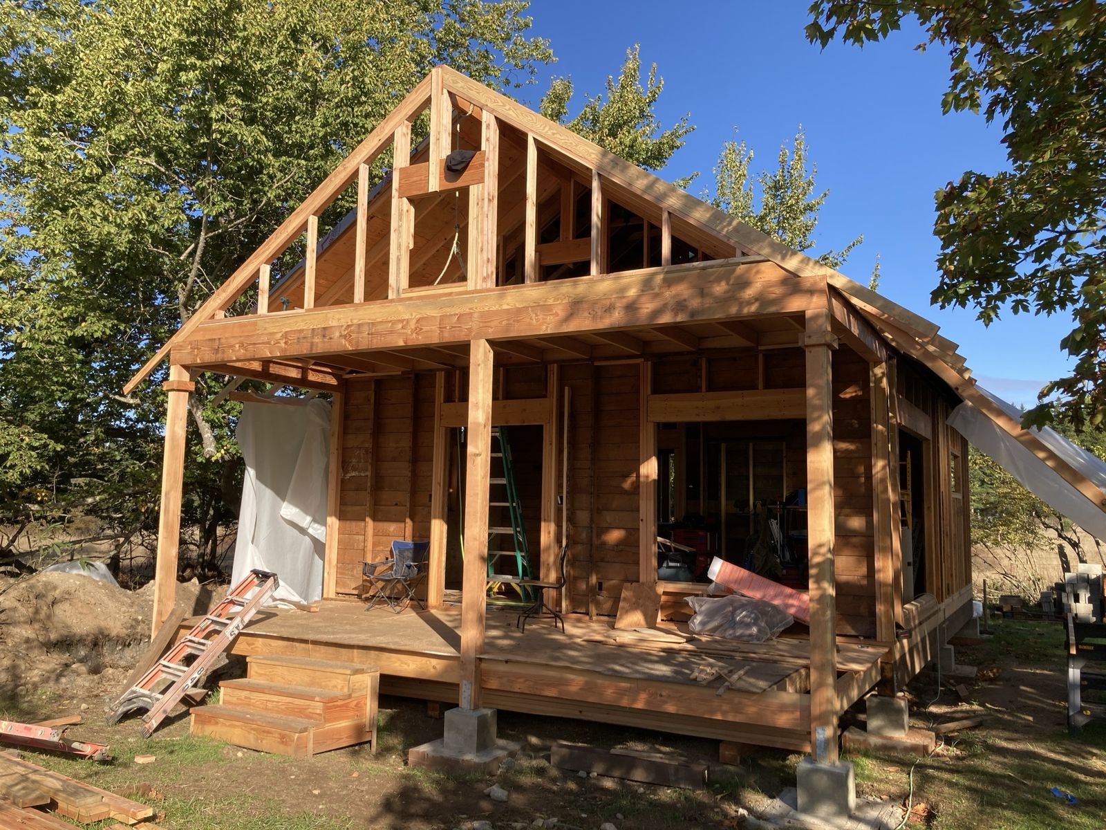 A house framed up in new timber against a blue sky, a steep gable over a covered porch, rafters and studs bare with no siding yet. A green stepladder and an orange one, a camp chair, a roll of red flashing and tools on the porch deck. Concrete piers underneath, maples in leaf on both sides.