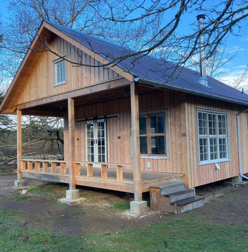 The finished house in winter — vertical cedar siding, a dark standing-seam roof, a covered porch with square posts and a low rail, a metal chimney flue running up the far side. Multi-pane windows, a glazed double door, wooden steps down to the grass. Bare branches overhead, blue sky.
