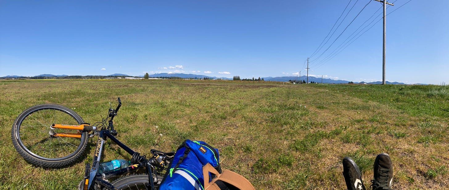 A bike laid down in a wide mown field, panoramic. Blue sky, a line of low mountains along the horizon, a power pole and its wires at the right. In the foreground the bike's front wheel, a blue dry bag strapped to the rack, a water bottle, and my own boots stretched out in the grass.