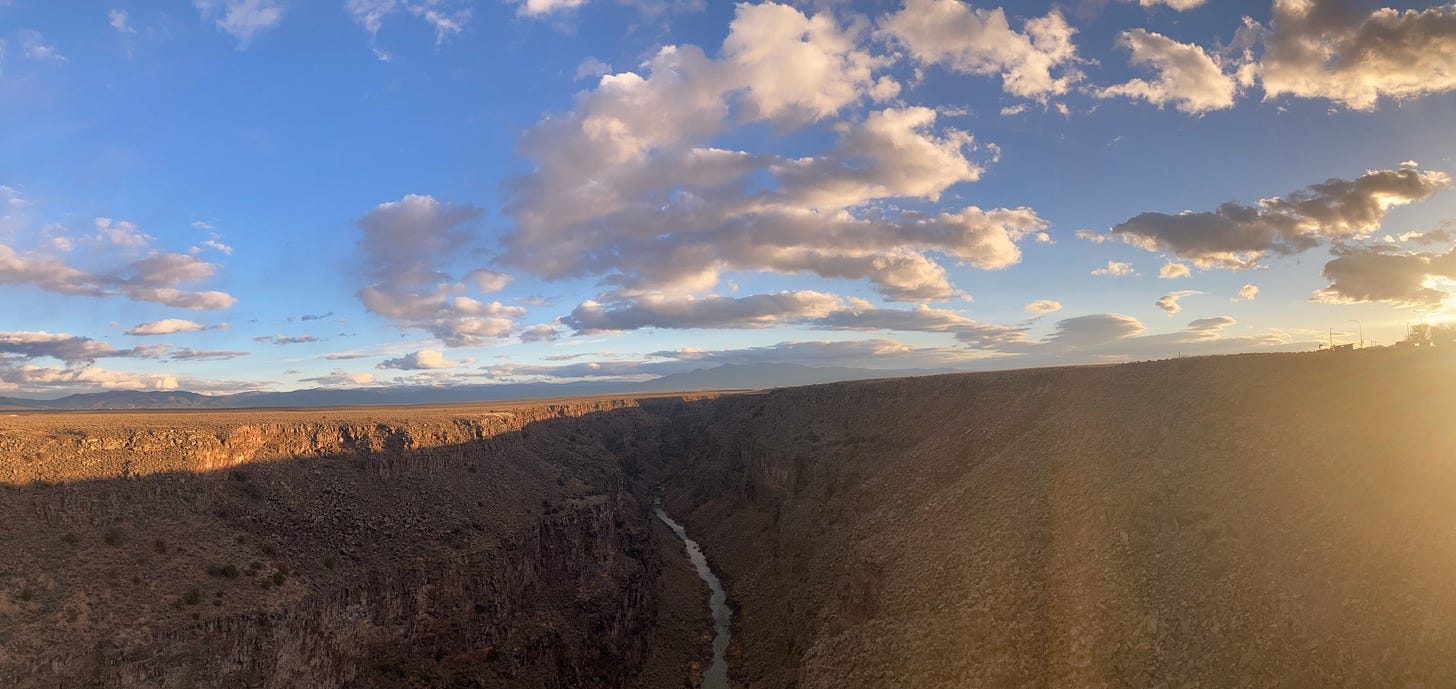 The Rio Grande gorge at sunset, panoramic. A dark basalt canyon splits flat desert, the river a thin band of light at the bottom. Long low clouds catch orange along the horizon, and the sun flares in from the right edge.