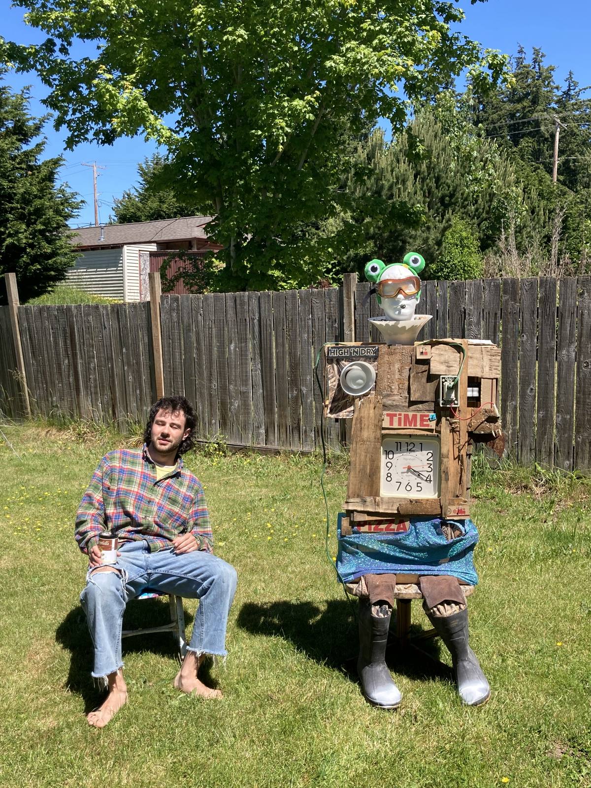 Soup sitting on a stool in a mown backyard against a wooden fence: a mannequin head in ski goggles and a green frog hat, a torso of pallet wood with a square wall clock for a belly and a speaker at the shoulder, a blue sequinned skirt, and grey rubber boots. Jack sits on a stool beside it, barefoot in a plaid shirt, holding a can.