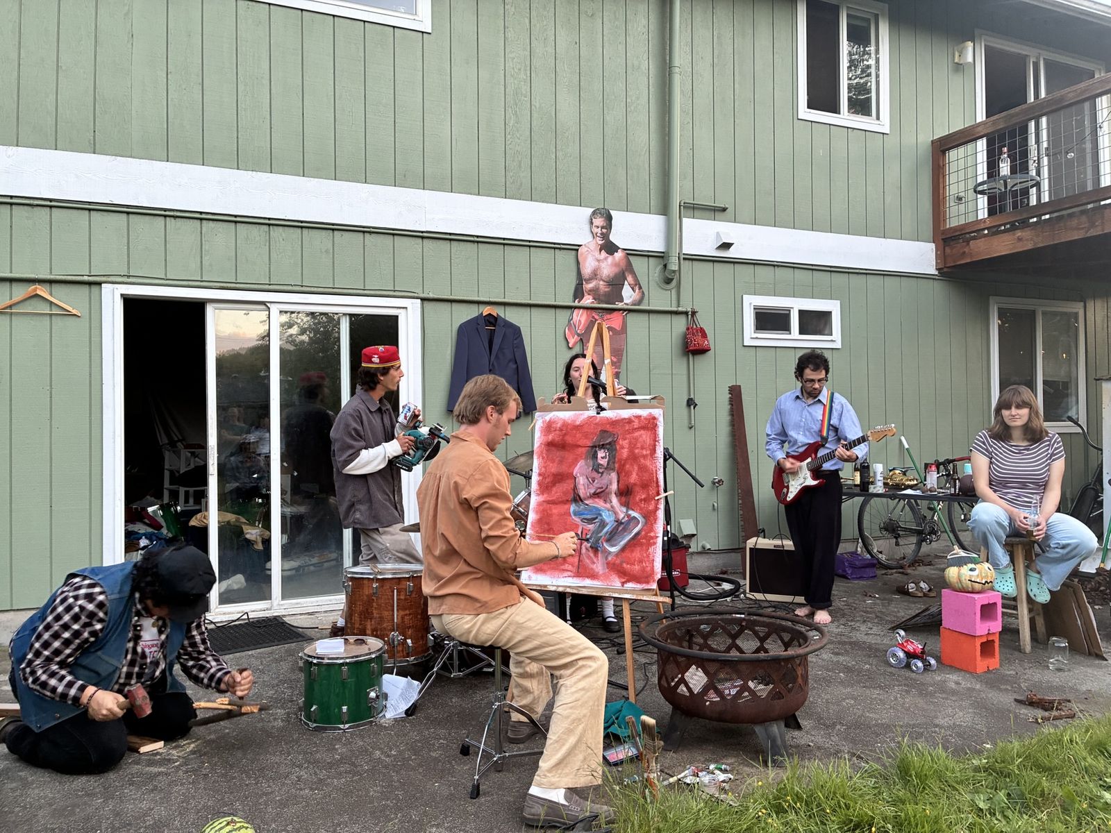 A performance underway on the concrete behind a green board-and-batten house. At the left a person in a denim vest and plaid shirt kneels on the ground working a chisel into a board. Behind them a drum kit, and a man in a red fez holding a camcorder. In the middle a man in a tan shirt sits at an easel, painting a seated figure onto a red canvas, with a singer at a microphone just behind it. At the right a man in a blue shirt and rainbow suspenders plays a red Stratocaster barefoot through a small amp, and a woman watches from a stool with a bike leaning behind her. A life-size cutout of a shirtless man is fixed to the wall above, beside a navy suit jacket on a hanger. A rusted fire pit stands in the middle of it all.