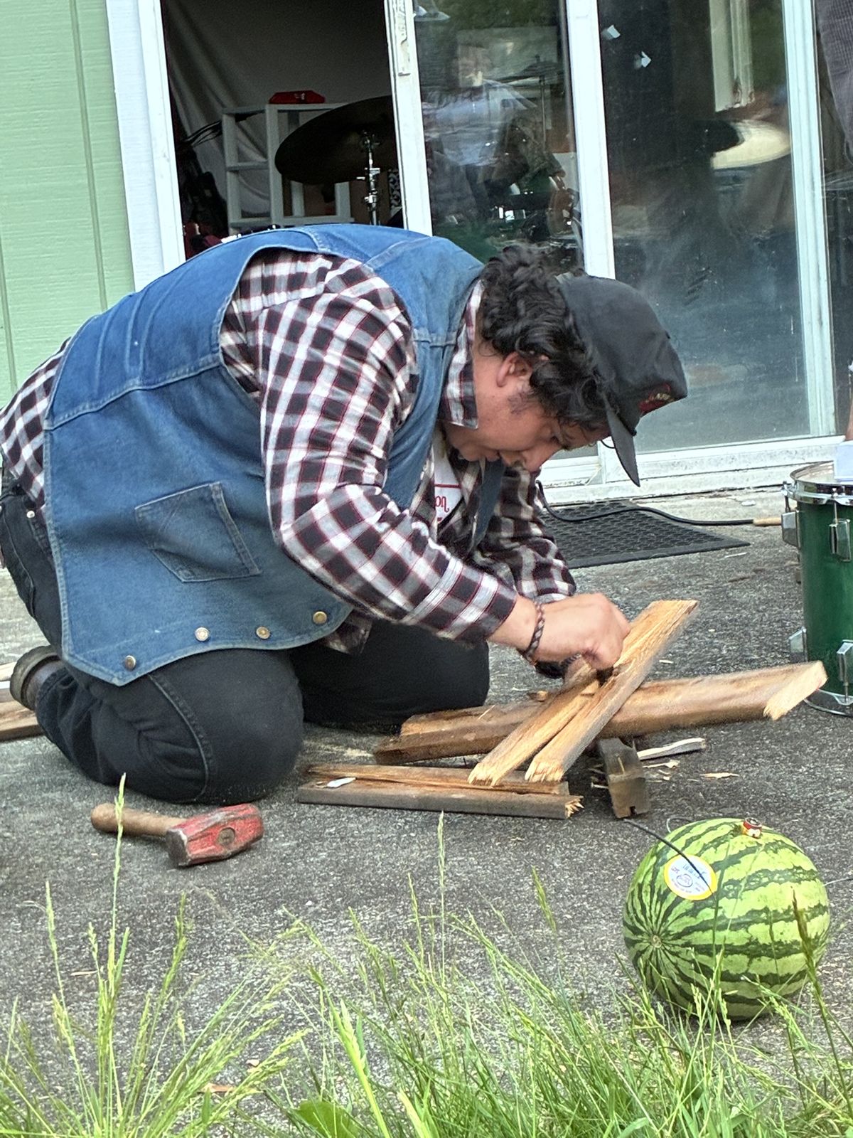 Closer on the same kneeling figure — denim vest over a red and white plaid shirt, black cap, dark curls — driving a chisel into a length of split fir. A red mallet lies on the concrete beside broken boards and a wooden mallet, a green drum at the edge of the frame, and a small watermelon with a wire taped into its top sits in the grass.