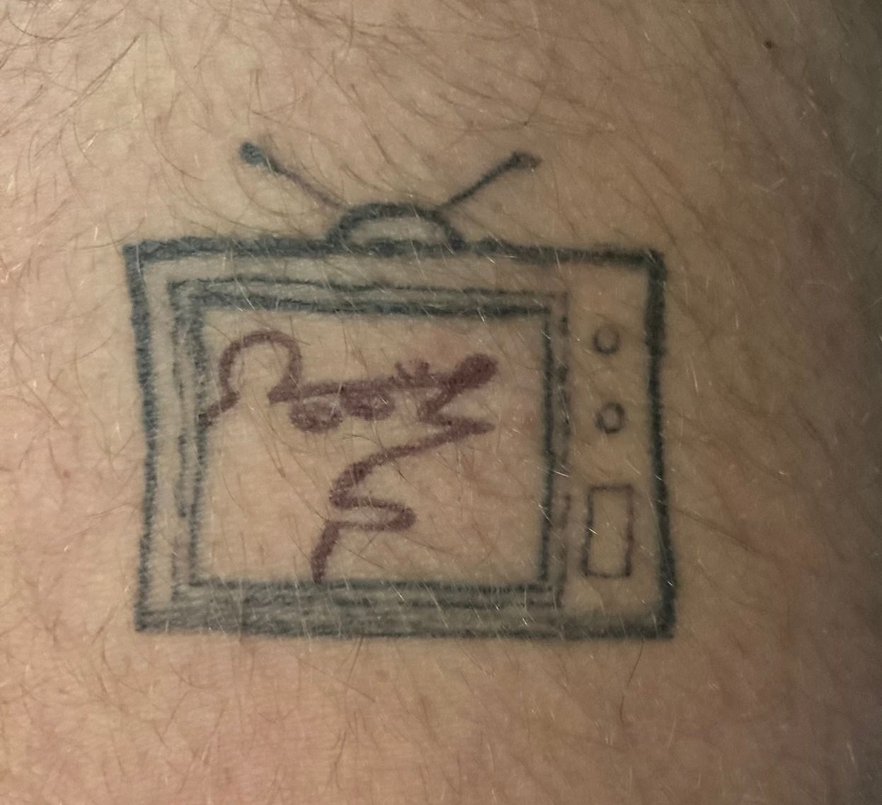 A small tattoo of an old television on hairy skin, drawn in black line: two rabbit-ear antennas, a double-lined screen, two dials and a slot down the right side. On the screen someone has drawn a loose red-ink squiggle.