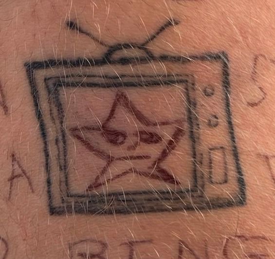The same television tattoo, closer. On its screen a star has been drawn in red pen with a sleepy little face in it, and more red handwriting runs around the skin outside the frame, too cut off to read.