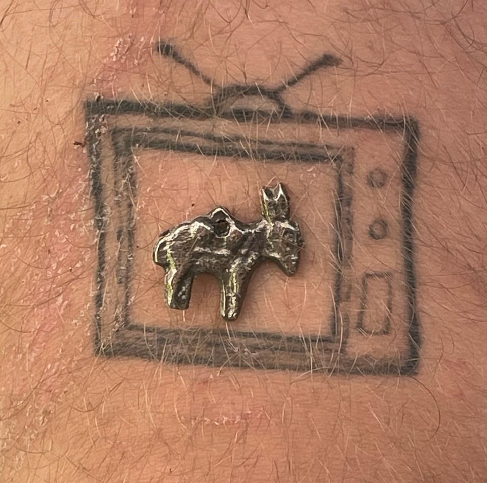 The television tattoo with a tiny cast-metal donkey charm, a hole through its back for a string, set down in the middle of the screen.